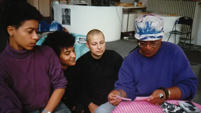 Four black people sit around a table as Audre Lorde looks through printed photographs. She wears a tie-dye hat and glasses. The others look closely at the photos.