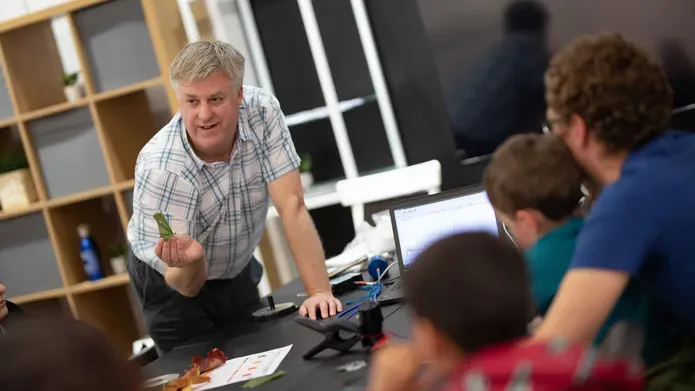 Man crouched over a table and laptop holding up a leaf and looking towards a man and two small children, who have their backs to us