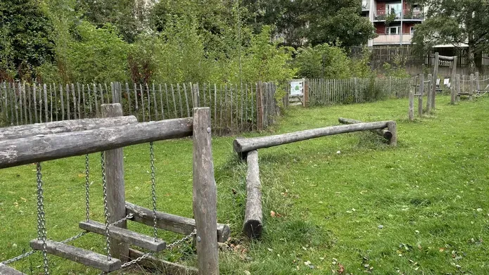 Kids wooden playframe and climbing equipment in grassy are with trees surrounding and tall buildings behind 