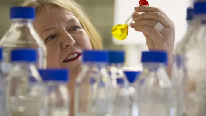 Close up image of woman holding up a small glass bottle with yellow liquid in it. In front of her are lots of glass jars with blue lids. 