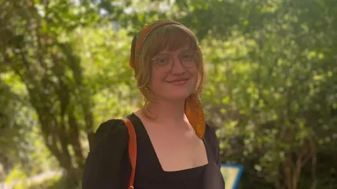 woman standing in forest looks towards camera and smiles. She is wearing a black top with a read bag strap over her shoulder, glasses and has fair hair tied back and a fringe 