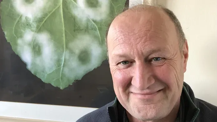 Head and shoulders shot of a man smiling. He is in front of a framed image of underside of green heart shaped leaf which has 4 fluffy circles, two on each side of the leaf.
