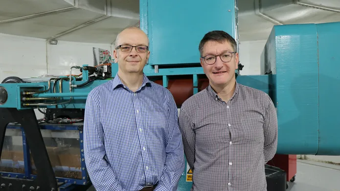 two men wearing checked shirts and glasses, looking and smiling at camera, with large machinery behind them