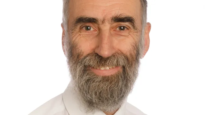 head and shoulders image of man in a white shirt and tie with grey beard, looking and smiling at camera. 