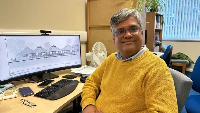 man wearing yellow woollen jumper and glasses, looking and smiling at camera. He is sat next to desk with a computer at it and a sketch of a building on the screen