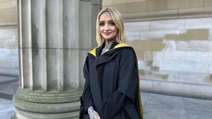 Young woman with long, straight blond hair and tattoos wearing a graduation gown, looking and smiling at camera