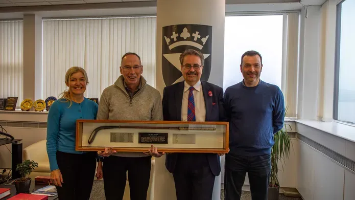 Four people stand smiling, holding a trophy case containing a putter and 3 plaques with names engraved.