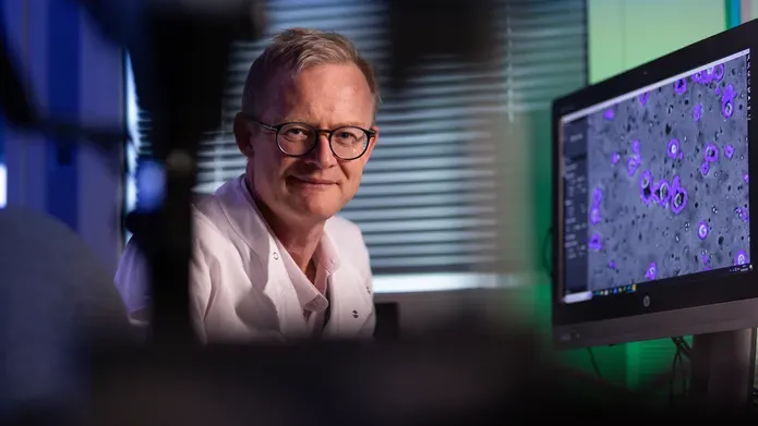 Man wearing glasses and whit lab coat looking towards the camera. Next to him is a screen with science cells on it. the image features blue and green lighting