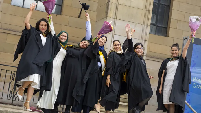 Seven Afghan women stand on the steps of a building wearing graduation robes and celebrating.