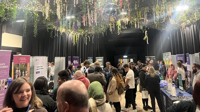 A crowd of people are in a room with flowers hanging from the ceiling, there are tables with banners promoting employers.