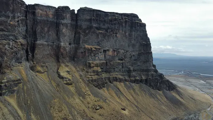 Aerial view of a steep cliff with layered rock formations and a flat landscape stretching into the distance under a cloudy sky.