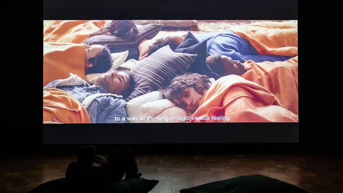 Person in a darkened room watching a large projection on a bean bag. The screen is the size of a double decker bus and shows a circle of people lying together on orange bean bags