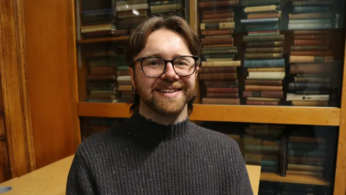 man with mid length dark hair sat in front of bookcase filled with books, looking and smiling at camera