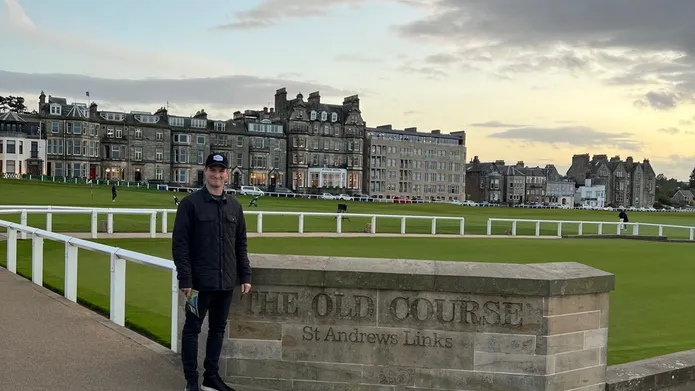 A man stands in front of a wall at the Old Course golf course, in St Andrews, there is a big stone building in the background.