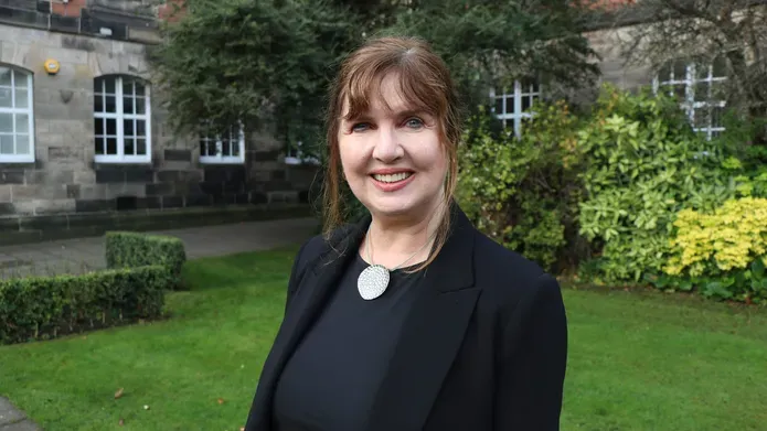 Woman with tied back dark hair, waring black smart outfit, looking and smiling at camera. She is outside in a garden area, with green bushes and grass behind her and old buildings