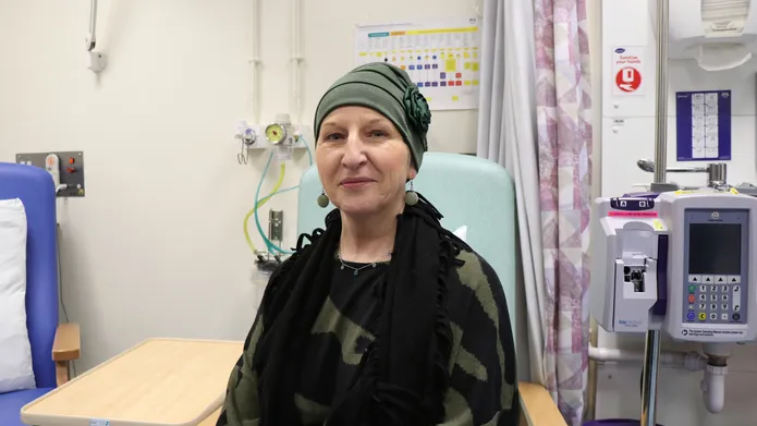 woman sat upright in chair, with green top and hat on, looking and smiling at camera. She is in a hospital room with some medical equipment around her