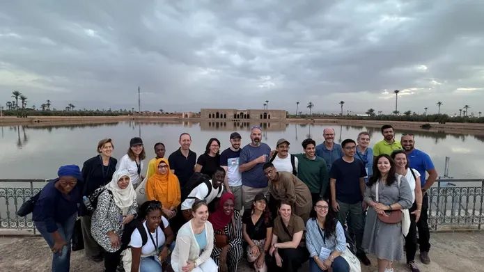 A group of people pose in front of a body of water with a building on the opposite shore.