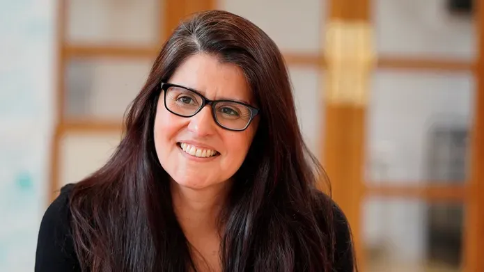 Close up head and shoulders shot of woman with long dark hair and glasses, looking slightly off camera and smiling