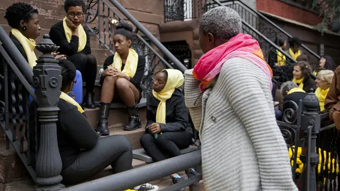 A group of women sit on the steps of a New York flat talking. A passerby stands and listens. The group talking wear yellow scarves