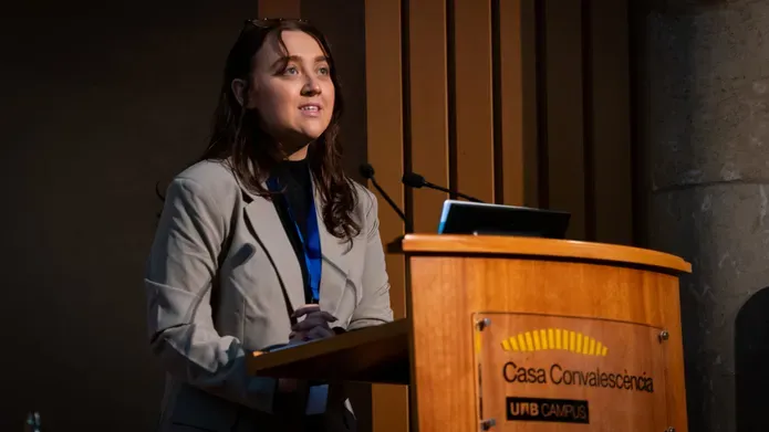 A girl with brown hair, wearing a grey blazer stands at a podium looking out to the top right.