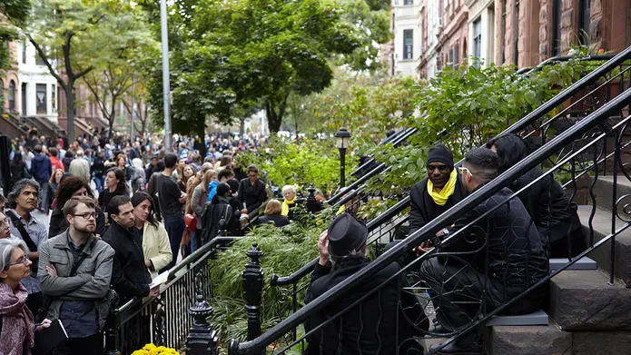 large crowd gather on a new york street, on a stoop are a group of women sitting and talking wearing yellow scarfs