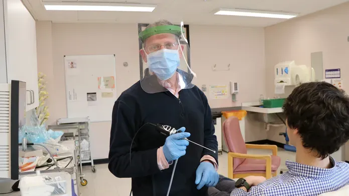man wearing a face mask and protective clear facial visor, holding a long thin tool with a light on the end of it. He is looking towards the camera. A patient is seen from behind lying on a hospital bed.