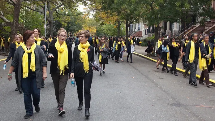 Many women walk down a Brooklyn street wearing yellow scarves, two women at the front are walking and talk