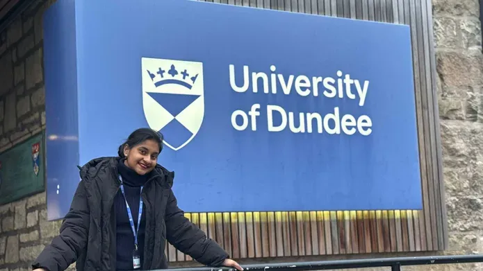 A woman in black clothing leans against metal railing outside of a building, there is a large blue 'University of Dundee' sign behind her.
