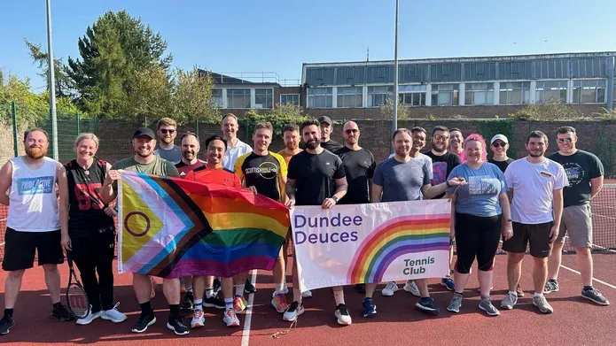 A crowd of people stand on an outdoor tennis court, holding LGBTQ+ and the Dundee Deuces flag.