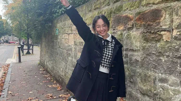 A girl in a black coat stands next to a tall stone wall with her right hand in the air doing the peace sign. 