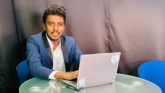 A man in a blue suit jacket sits at a table with a laptop in front of him.
