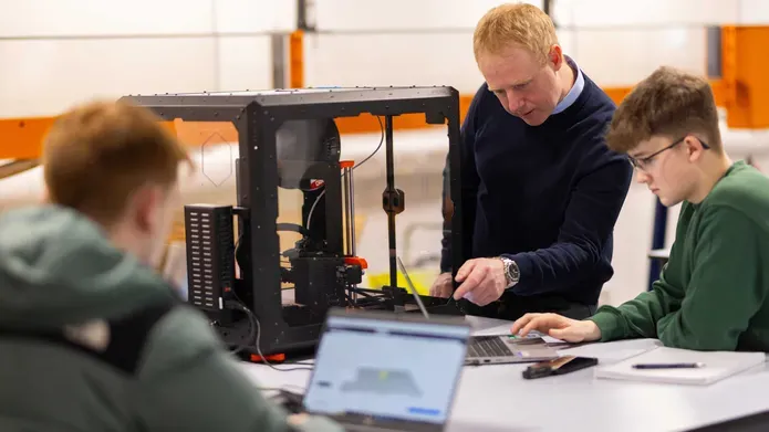 Two students work on laptops next to a 3d printing machine, a tutor stands next to one of the students pointing at a laptop screen.