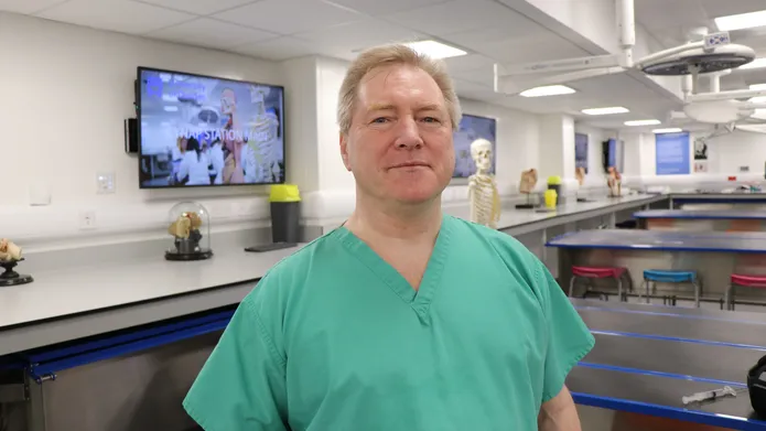 man wearing hospital scrubs looking at camera and smiling. Behind him are tv screens on the walls with medical image on them