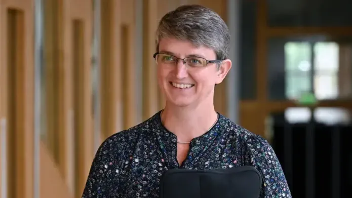 A woman in a dark blouse with short hair smiling in walking down a hallway