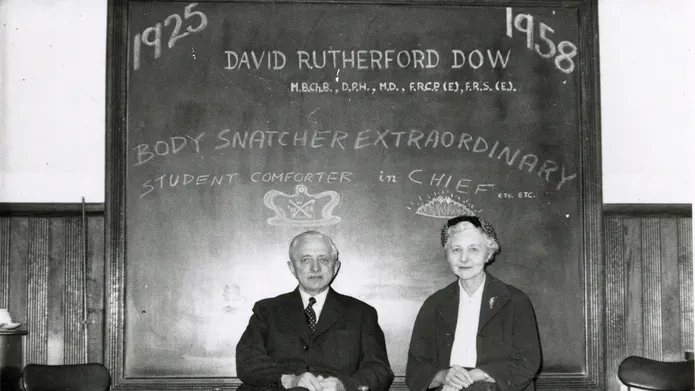 A black and white photo of Professor David Dow and Dr Agnes Dow sitting in front of a blackboard. The board reads 1925 1958 David Rutherford Dow Body Snatcher Extraordinary Student Comforter in Chief etc., etc. Above David's head is a crown drawn on the blackboard, above Agnes's, a tiara.