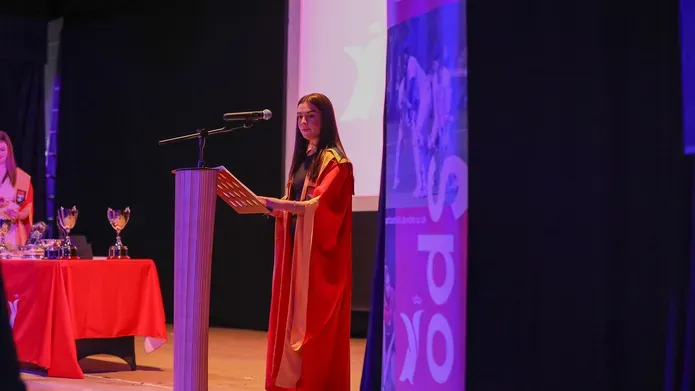 A person in a robe stands in front of a pedestal speaking in a microphone.