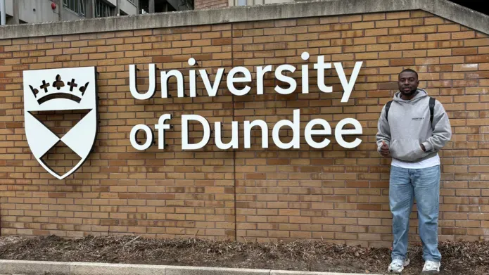 A man stands in front of a brick wall with the University of Dundee written on it and the UoD shield.