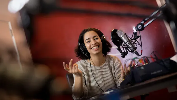 A person sits at a microphone wearing a headset and smiling with their hands raised in front of them