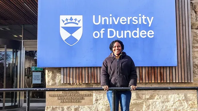 A woman stands with her hands on black metal railing, in front of a blue University of Dundee sign.