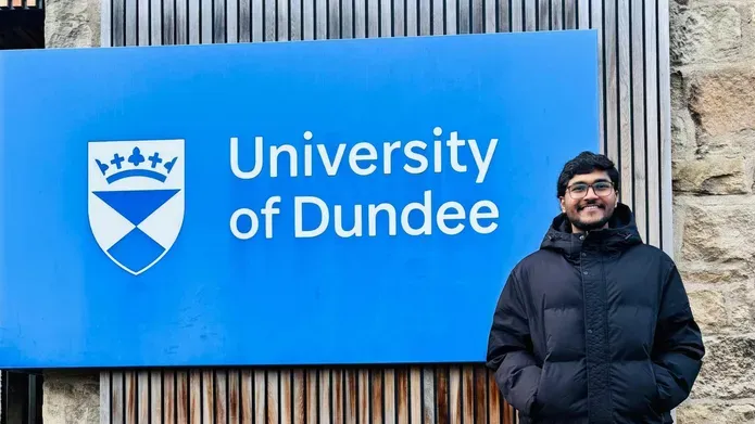 A person stands in front of a large blue University of Dundee sign, with his hands in his jacket pockets, smiling.