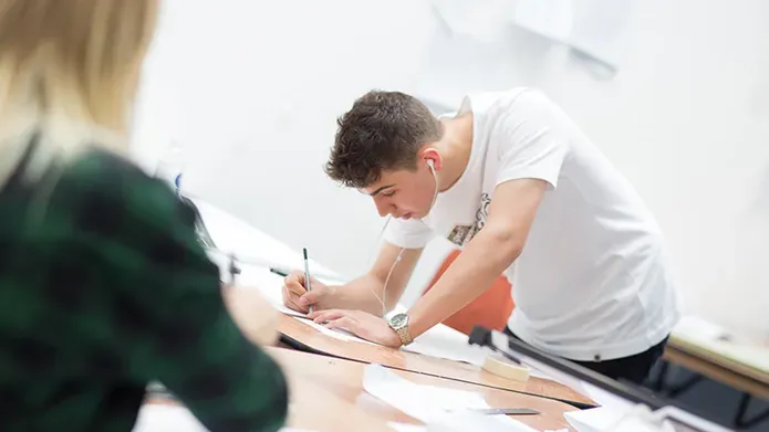 A male in a white t-shirt leans over a desk with a pencil in his hand, drawing.