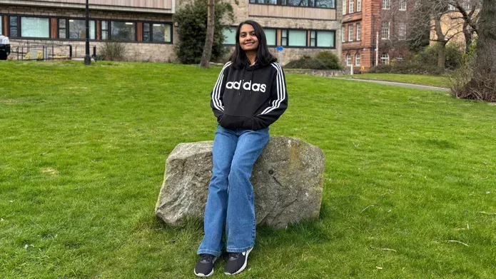 A person sits on a large boulder wearing a black hoodie and blue jeans, with their hands in their pockets.