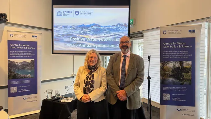 A man and woman stood smiling in front of a projector screen and 2 banners