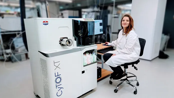 A person in lab coat sat on a chair in front of a desktop computer. Attached to the computer is a large box approx. 1.5m high which contains the scientific instrument.