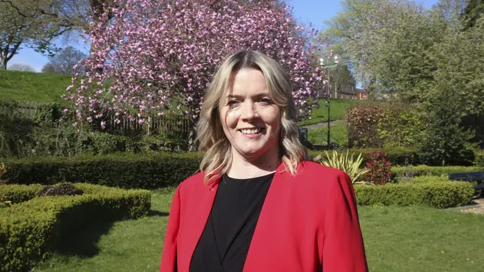 Kirsty Thomson wearing a bright red blazer, standing in a well tended garden with a pink blossoming tree behind her.