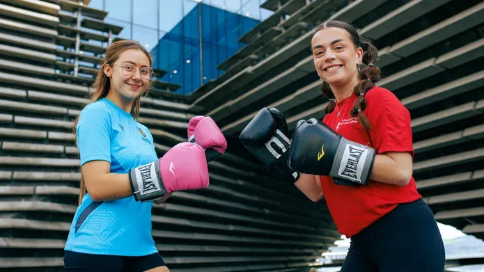 Two people stand in front of the V&A building holding their hands up to each other, wearing boxing gloves.