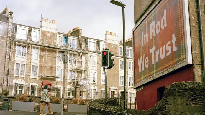A billboard with a tartan background with "In Rod we Trust" written in large white letters on a Dundee street.