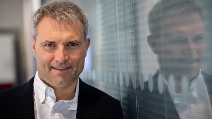Man in white shirt and dark jacket looking at camera with his reflection seen in the glass wall on his left.