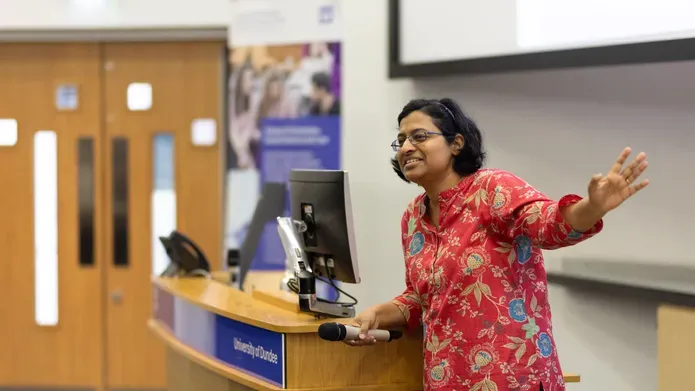 Dr Edzia Carvalho addresses visitors, standing infront of a podium in a lecture theatre.