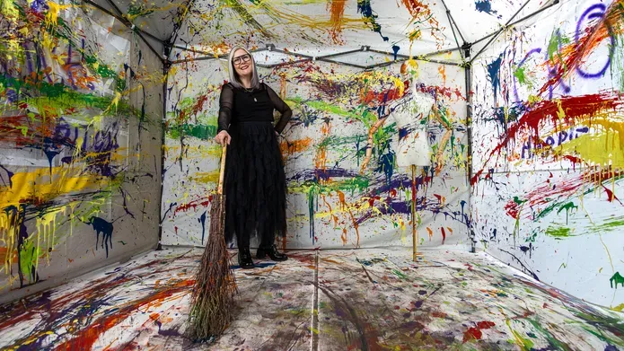 Nicky standing in her installation, holding a witches' broom. The walls are cover in different colours of paint and a mannequin is in the foreground, also covered in differnet colours.
